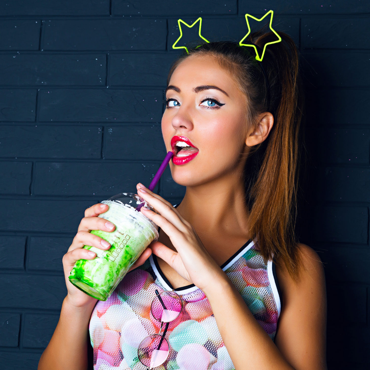 Urban fashion portrait of sexy brunette girl with hight ponytail, trendy make up, wearing printed shirt and funny party stars accessory on her head, drinking tasty sweet green milkshake , urban style.